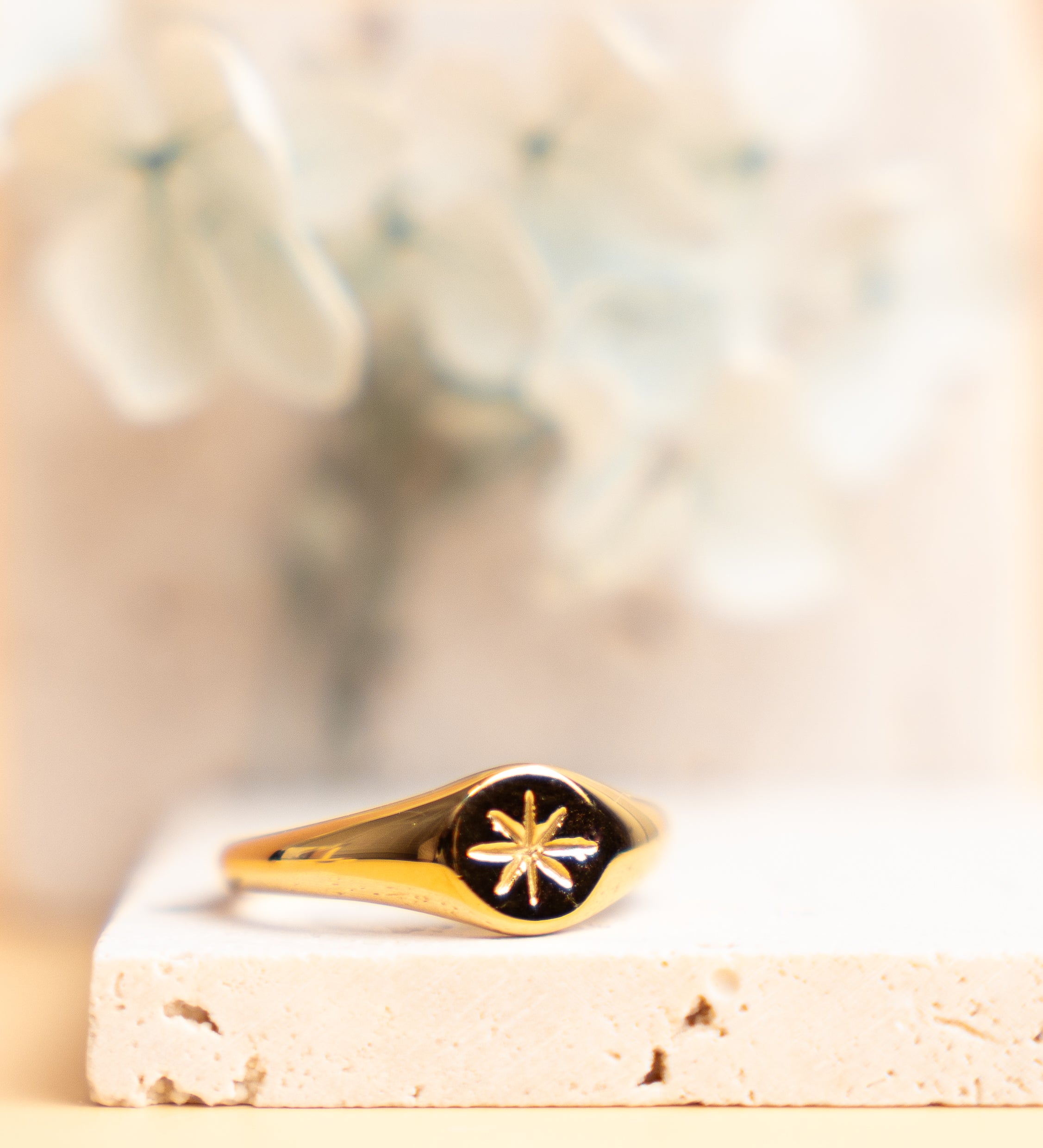 An 18K gold plated signet ring with a star-shaped detail on the top, placed on a white surface with a blurred background.